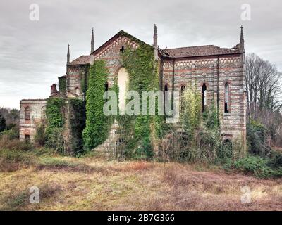 Alte verlassene Steinkirche überwuchert mit Unkraut und Kriechen, die von einem angrenzenden Feld unter einem bewölkten Himmel betrachtet werden Stockfoto