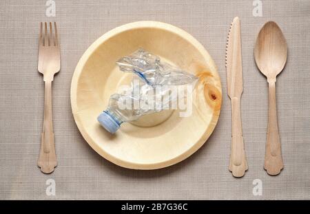 Kunststoffkonzept mit Tischeinstellung und zerknitterter Kunststoffflasche auf einer natürlichen Holzplatte, die von passendem Messer, Gabel und Löffel aus gesehen flankiert wird Stockfoto