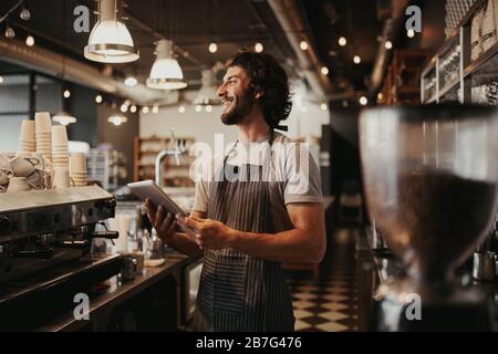 Ein gutaussehender kaukasischer Mann im Café lacht, während er das digitale Tablett in der Hand hinter der Theke hält Stockfoto