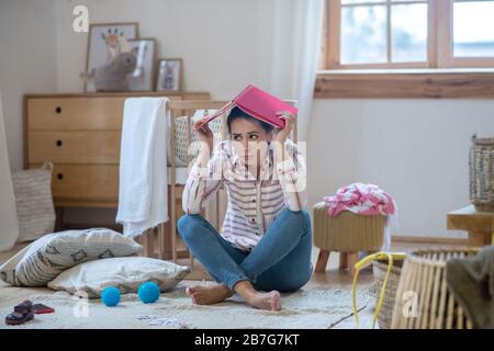 Müde Frau sitzt auf dem Boden mit Notizbuch auf dem Kopf Stockfoto