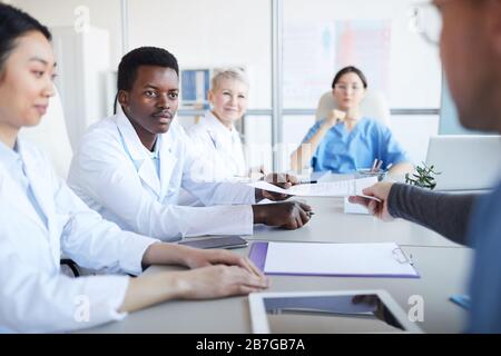 Portrait des jungen afroamerikanischen Arztes, der während einer medizinischen Konferenz mit Kollegen am Tisch Vertrag oder Dokument ergreift, Kopierraum Stockfoto