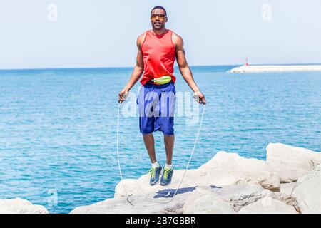 Schwarzer gutaussehender Mann mit Springseil mit Meerblick im Hintergrund - Außenläufer, der Sport macht und sich auf das morgendliche Workout vorbereitet - gesunder Lebensstil Stockfoto