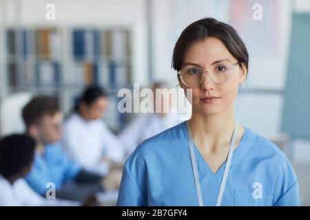 Kopf- und Schulterporträt junger Krankenschwestern, die eine Brille tragen und die Kamera vor dem Hintergrund medizinischer Konferenzen betrachten Stockfoto