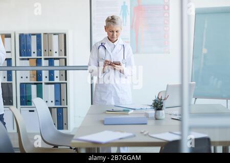 Portrait eines reifen Hausarztes, der Smartphone verwendet, während er im Konferenzraum im Inneren des medizinischen Büros am Tisch steht, Kopierraum Stockfoto