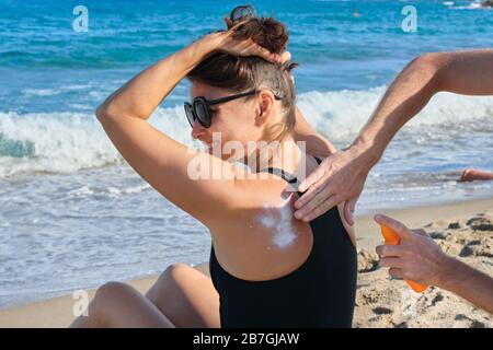 Frau mittleren Alters am Strand, männliche Hände mit sonnenförmiger Creme Stockfoto