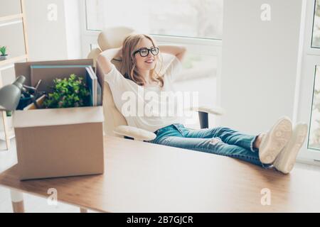 Konzept der Förderung bei der Arbeit. Junge süße lächelnde Frau, die mit ihren Füßen auf dem Tisch im Sessel im neuen Büro sitzt und sich ausruhen kann Stockfoto