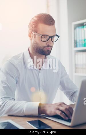 Selbstbewusster bärtiger Geschäftsmann mit Brille und weißem Hemd funktioniert auf dem Laptop im Büro Stockfoto