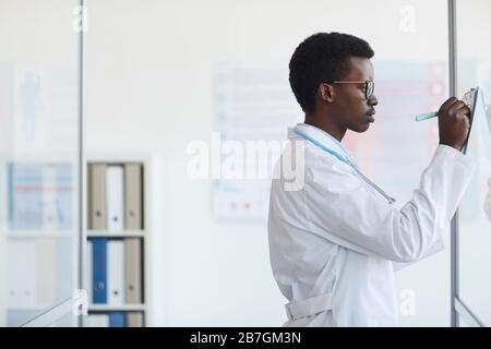 Seitenansicht Porträt des jungen afroamerikanischen Arztes, der in der Zwischenablage schreibt, während er an der Glaswand in der Klinik steht, Kopierraum Stockfoto