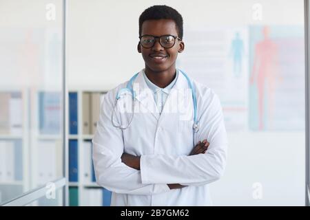 Taille oben Porträt des jungen afroamerikanischen Arztes, der mit den Armen gekreuzt und lächelnd auf der Kamera steht, während er in der Klinik posiert, kopiere Raum Stockfoto