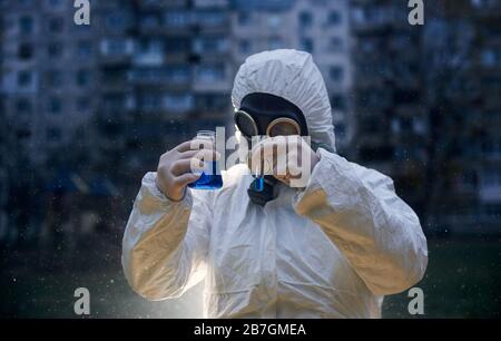 Wissenschaftler, die einen Overall und eine Gasmaske tragen und Glaskolben mit blauem Reagenz vor einem hochgeschossigen Schnappschuss aus der Nähe halten Stockfoto