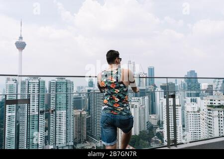 Mann mit Sonnenbrille und Sommerkleidung auf dem Rücken, der über eine Stadt in Kuala Lumpur in Malaysia nachdenkt Stockfoto