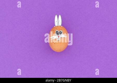 Braunes osterei mit lustigen Hünenohren und Cartoon-Gesicht auf violettem Hintergrund Stockfoto