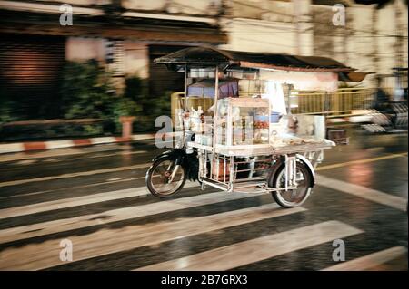 Thailändisches Straßennahrungsmittelgeschäft, das nachts in Phuket in der Regenzeit mit dem Dreirad fährt. Hohe iso-empfindlichkeit, Bewegungsunschärfe und Körnung. Stockfoto