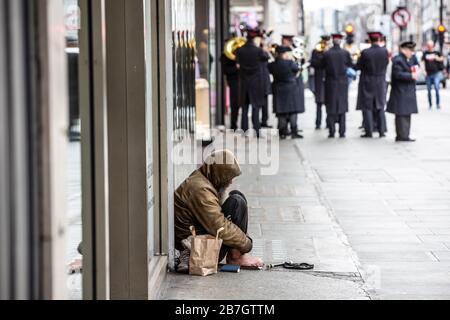 Obdachloser sitzt an einem Schaufenster, während die Heilsarmee Band in der Ferne Passanten in der Oxford Street, London, UK, Musik spielt Stockfoto