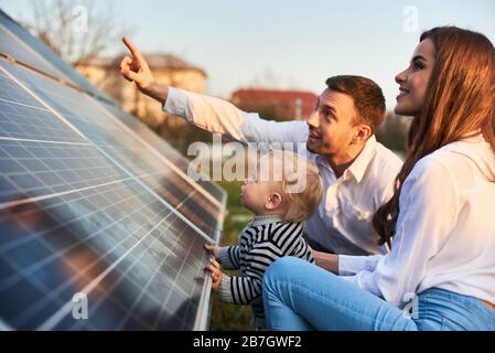 Der Mann zeigt seiner Familie die Sonnenkollektoren auf dem Grundstück in der Nähe des Hauses während eines warmen Tages. Junge Frau mit Kind und Mann in den Sonnenstrahlen sehen sich die Sonnenkollektoren an. Stockfoto