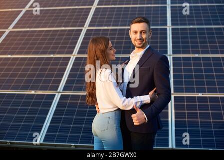 Wunderschönes Paar, das sich auf dem Hintergrund von Solarpaneelen umarmt. Schlankes Mädchen in Jeans mit langem Haar sieht mit Bewunderung auf einen Kerl in einer Jacke aus, der wie ein Daumenschild zeigt. Bild des Solarenergiekonzepts Stockfoto