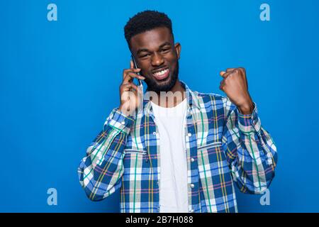 Junger afrikanischer Mann, der ein Telefon hält, jubelte sorglos und freute sich über den blauen Hintergrund Stockfoto