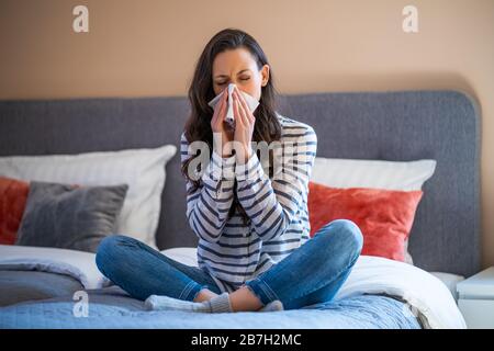 Frau sitzt in ihrem Haus auf dem Bett, sie hat Grippe und bläst Nase. Stockfoto