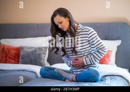 Frau sitzt auf dem Bett in ihrem Haus, sie hat Schmerzen im Magen. Stockfoto