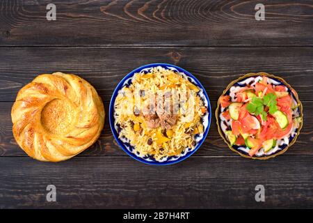 Nationaler usbekischer Pilaf mit Fleisch, Achichuk-Salat aus Tomaten, Gurke, Zwiebel in Platte mit traditionellem Muster, Cilantro, Kirschtomaten, Garlikbrot Stockfoto
