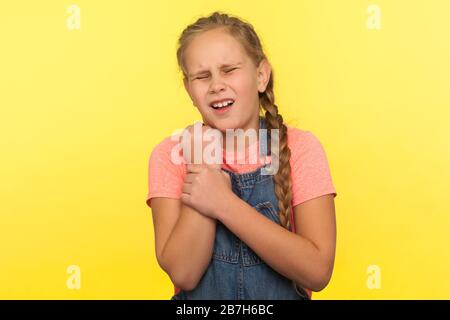 Kind mit verletzter Hand. Porträt des verärgerten kleinen Mädchens in Denim-Overalls, das mit Schmerzen und dem Berühren des Wunde gespreizten Handgelenks steht, medizinische Probl Stockfoto