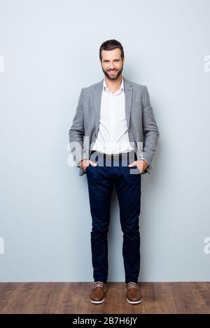 Vertikales, durchgehaltener Portrait eines gutaussehenden lächelnden, glücklichen und erfolgreichen Geschäftsmannes, der seine Hände in den Taschen auf grauem Hintergrund hält Stockfoto