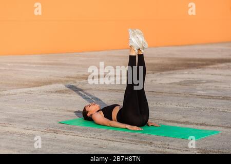 Passen Sie die schlanke junge Frau in enge Sportswear, schwarze Hosen und Oberteil, liegen auf der Matte, halten Sie die geraden Beine hoch, trainieren Sie Bauchmuskeln, Flexibilität. Hea Stockfoto