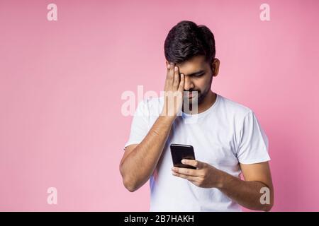 Nahaufnahme des attraktiven jungen jungen, unrasierten indischen Männchens, das weißes T-Shirt trägt, Smartphone hält, auf den Bildschirm blickt, mit Hand ein Auge bedeckt, Isola Stockfoto