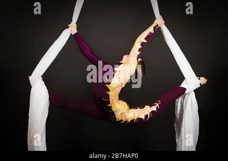 Sportliche Frau im Burgunder Anzug führt Gymnastik- und Zirkusübungen auf weißer Seide durch. Studio-Aufnahmen vor dunklem Hintergrund, isolierte Bilder. Stockfoto