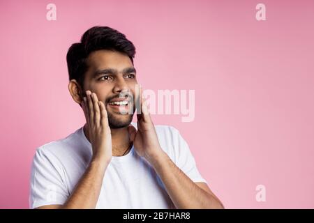 Porträt eines angenehm überraschten glücklichen Mannes in weißem T-Shirt, Berühren von Wangen mit Handflächen, beiseite mit verblüfftem Ausdruck, isoliert auf pinkfarbenem Backgr Stockfoto