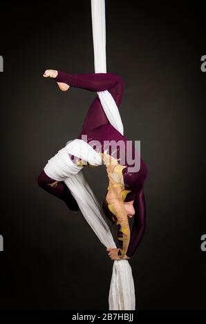 Sportliche Frau im Burgunder Anzug führt Gymnastik- und Zirkusübungen auf weißer Seide durch. Studio-Aufnahmen vor dunklem Hintergrund, isolierte Bilder. Stockfoto