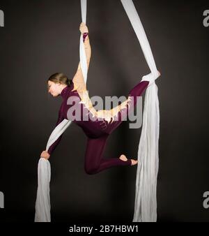 Sportliche Frau im Burgunder Anzug führt Gymnastik- und Zirkusübungen auf weißer Seide durch. Studio-Aufnahmen vor dunklem Hintergrund, isolierte Bilder. Stockfoto