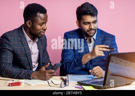 Ein gutaussehender junger afroamerikanischer Geschäftsmann, der sein Projekt auf einem Laptop vor einem indischen Geschäftspartner präsentiert und in Offi am Tisch sitzt Stockfoto