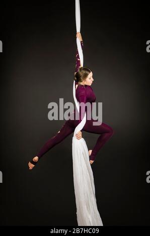 Sportliche Frau im Burgunder Anzug führt Gymnastik- und Zirkusübungen auf weißer Seide durch. Studio-Aufnahmen vor dunklem Hintergrund, isolierte Bilder. Stockfoto