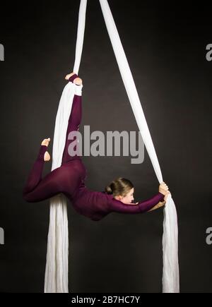 Sportliche Frau im Burgunder Anzug führt Gymnastik- und Zirkusübungen auf weißer Seide durch. Studio-Aufnahmen vor dunklem Hintergrund, isolierte Bilder. Stockfoto