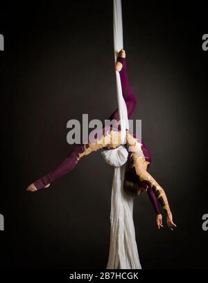 Sportliche Frau im Burgunder Anzug führt Gymnastik- und Zirkusübungen auf weißer Seide durch. Studio-Aufnahmen vor dunklem Hintergrund, isolierte Bilder. Stockfoto