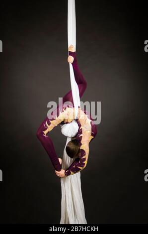 Sportliche Frau im Burgunder Anzug führt Gymnastik- und Zirkusübungen auf weißer Seide durch. Studio-Aufnahmen vor dunklem Hintergrund, isolierte Bilder. Stockfoto