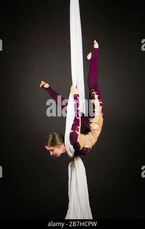 Sportliche Frau im Burgunder Anzug führt Gymnastik- und Zirkusübungen auf weißer Seide durch. Studio-Aufnahmen vor dunklem Hintergrund, isolierte Bilder. Stockfoto