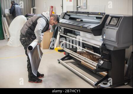 Der Bediener des Technikers wechselt die Papierrolle auf einem großen hochwertigen Industriedrucker und Plottergerät im digitalen Druckereibüro Stockfoto