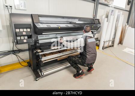 Der Bediener des Technikers wechselt die Papierrolle auf einem großen hochwertigen Industriedrucker und Plottergerät im digitalen Druckereibüro Stockfoto