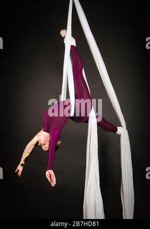 Sportliche Frau im Burgunder Anzug führt Gymnastik- und Zirkusübungen auf weißer Seide durch. Studio-Aufnahmen vor dunklem Hintergrund, isolierte Bilder. Stockfoto