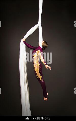 Sportliche Frau im Burgunder Anzug führt Gymnastik- und Zirkusübungen auf weißer Seide durch. Studio-Aufnahmen vor dunklem Hintergrund, isolierte Bilder. Stockfoto