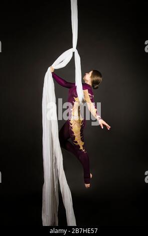 Sportliche Frau im Burgunder Anzug führt Gymnastik- und Zirkusübungen auf weißer Seide durch. Studio-Aufnahmen vor dunklem Hintergrund, isolierte Bilder. Stockfoto