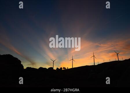 Dämmerungsstrahlen bei Sonnenuntergang über einer Windfarm, deren Silhouette bei Hintergrundbeleuchtung herausgeschnitten wird Stockfoto