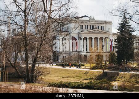 Das Gebäude der lettischen Nationaloper, das im Jahre 1863 im griechischen klassischen Stil erbaut wurde, durchzieht die Bäume, während an einem Winternachmittag Sonnenschein ausströmt Stockfoto