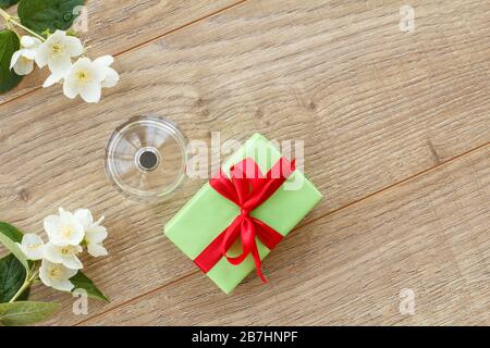 Kleine Geschenkbox mit rotem Band und einer Flasche Parfüm auf Holzbrettern, die mit Jasminblüten verziert sind. Draufsicht. Stockfoto