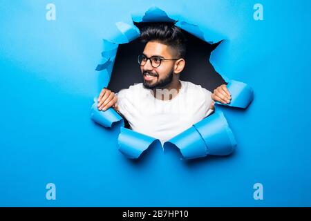 Der junge indische bärtige Mann sieht aus blauem Papierloch lächelnd aus Stockfoto