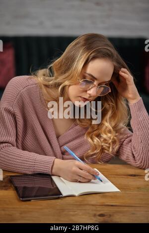 Blonde Bloggerin in pinkfarbener Strickjacke und Brille. Sitzt am Holztisch und schreibt etwas in ihr Notizbuch. Tablett liegt in der Nähe. Nahaufnahme Stockfoto