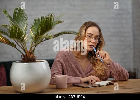 Mädchen Blogger in rosa Strickjacke und Brille. Am Holztisch sitzen und etwas in Notizbuch schreiben. Palmenbaum in Topf, Becher und Tablett in der Nähe Stockfoto