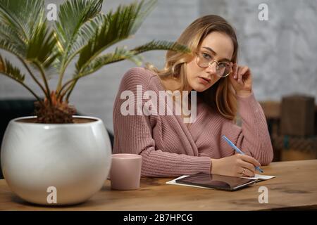 Frau in rosafarbener Strickjacke und Brille. Sitzt am Holztisch und schreibt etwas in ihr Notizbuch. Palmenbaum in Topf, Becher und Tablett. Nahaufnahme Stockfoto